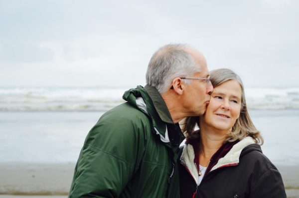 Ein älterer Mann und eine Frau stehen am Strand, der Mann gibt der Frau einen Kuss auf die Wange