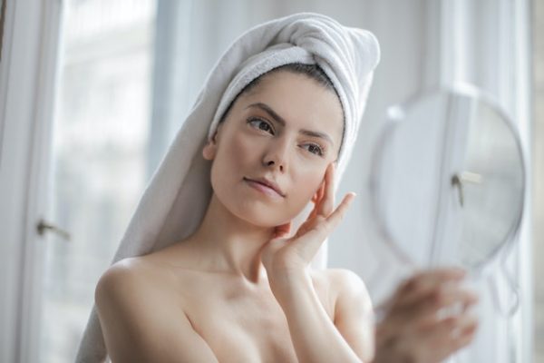 Eine Frau, mit in ein Handtuch eingewickelten Haaren, hält mit einer Hand einen Spiegel. Mit der anderen streicht sie ihre Wange