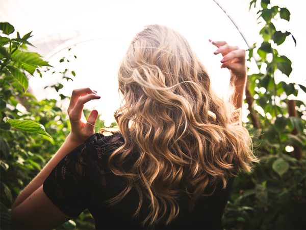 Eine Frau mit blonden lockigen Haaren steht mit dem Rücken zur Kamera vor einem Gebüsch und hebt die Hände in Richtung ihres Kopfes